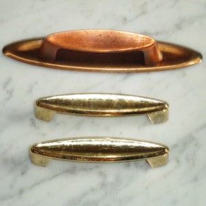 Puede incluir: Tres tiradores de armario, dos son dorados y uno es de cobre. El tirador de cobre tiene forma ovalada y los tiradores dorados son más rectangulares.