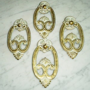 以下が含まれることがあります： 装飾的なデザインが施された、アンティークな白と金のキャビネットドアノブが4つ。ノブはリングハンドルで、楕円形です。