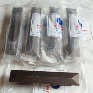 Puede incluir: Cinco paquetes individuales de tiradores de armario de bronce oscuro con forma cuadrada. Cada paquete tiene una etiqueta blanca con el texto "Hickory Hardware" y una dirección de sitio web.
