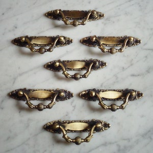 May include: Seven antique brass drawer pulls with ornate detailing. Each pull has a rectangular backplate, a curved handle, and decorative accents. The pulls are arranged on a marble surface, showcasing their vintage design.