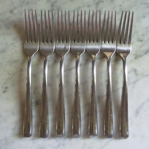 Puede incluir: Una colección de ocho tenedores de acero inoxidable dispuestos en fila. Cada tenedor tiene cuatro púas y un mango largo y delgado. Los tenedores se muestran sobre una superficie clara, similar al mármol.