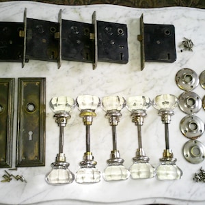 Puede incluir: Un surtido de herrajes antiguos para puertas, que incluyen varias cerraduras de embutir de metal negro, placas de puerta de latón y pomos de puerta de cristal transparente con vástagos de metal. También se incluyen rosetas de color latón y tornillos pequeños.