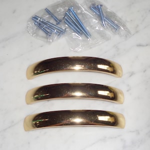 Puede incluir: Tres tiradores de puerta de armario de color dorado con forma curva. Los tiradores están dispuestos en fila, con el tirador superior ligeramente más alto que los demás. Los tiradores son de metal y tienen un acabado liso y pulido.