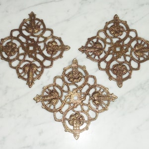 Op de afbeelding: Drie sierlijke, goudkleurige metalen decoratieve stukken met een vierkant gat in het midden. Elk stuk heeft een ruitvorm met ingewikkelde bloemmotieven.