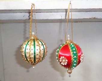May include: Two Christmas ornaments. One is cream-colored with green stripes and gold accents. The other is red with green and gold trim. Both have gold hanging cords and pearl-like embellishments.