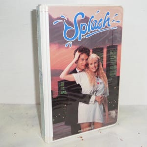 May include: A VHS tape of the film "Splash" with a couple in front of a city skyline. The title "Splash" is in blue script. The tape is in a white case.