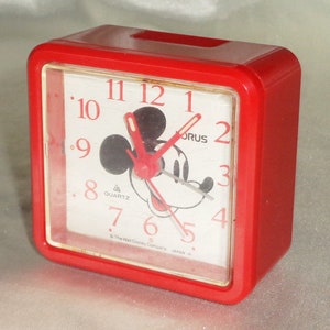 May include: Red plastic alarm clock with a Mickey Mouse face on the front. The clock face has black hands and black numbers. The clock says "Quartz" and "The Walt Disney Company Japan".