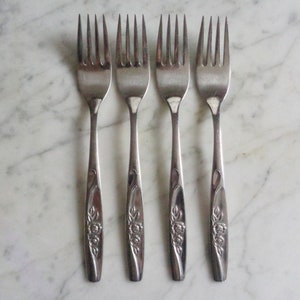 Pode incluir: Quatro garfos de prata com desenhos florais nas alças. Os garfos estão dispostos em uma fileira sobre uma superfície de mármore branco.
