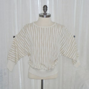May include: A white sweater with gold stripes and a ribbed hem. The sweater is on a white mannequin.