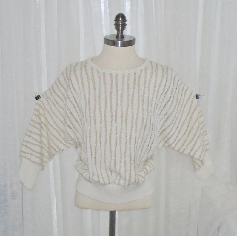 May include: A white sweater with gold stripes and a ribbed hem. The sweater is on a white mannequin.