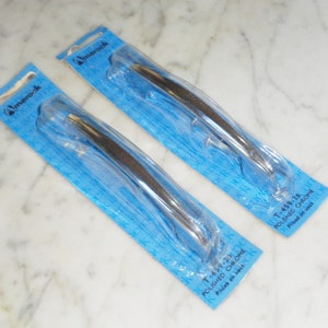 Puede incluir: Dos manillas de puerta plateadas envasadas en un embalaje de plástico azul. El embalaje tiene el texto "Amerock" y "Cromado pulido". El texto "T-459-28" y "T-459-23" también está en el embalaje.