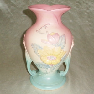 Puede incluir: Un jarrón de cerámica vintage con un esmalte rosa, blanco y azul. El jarrón presenta un diseño floral con flores amarillas, rosas y azules. El jarrón tiene dos asas y una base redondeada.