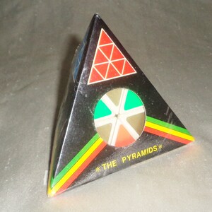 May include: A black pyramid-shaped puzzle toy featuring a red triangle with white grid lines, a circular cutout revealing internal colored segments, and the text THE PYRAMIDS printed in yellow near the base with green and red accent stripes.