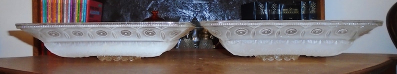 May include: Two white frosted glass bowls with a decorative pattern. The bowls are oval shaped and have a scalloped edge.