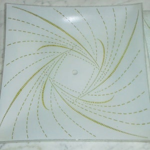 Pode incluir: Uma lumin&aacute;ria de teto quadrada branca com um padr&atilde;o de linhas douradas em espiral. As linhas s&atilde;o feitas de pequenos tra&ccedil;os e criam um efeito de espiral.