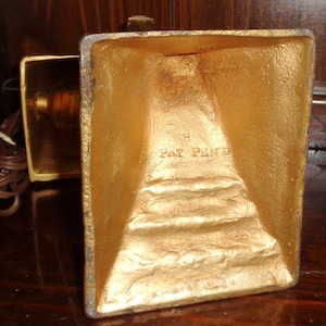 May include: Gold-colored metal lamp base with a square shape and a rectangular base. The base has a raised inscription that reads "Pat Pend".