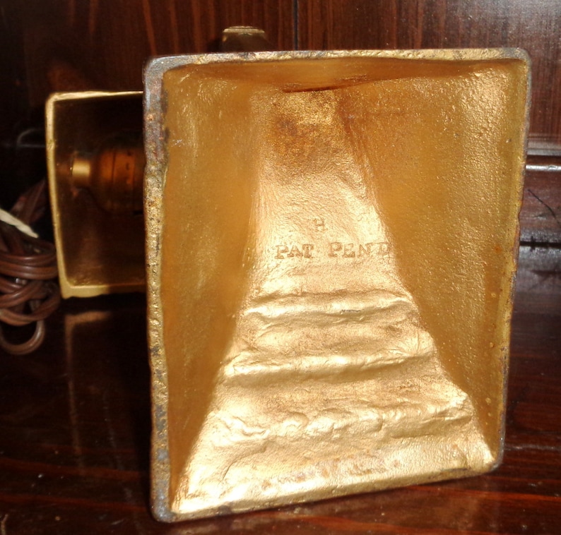 May include: Gold-colored metal lamp base with a square shape and a rectangular base. The base has a raised inscription that reads "Pat Pend".
