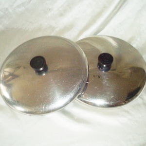 Puede incluir: Dos tapas de olla plateadas con pomos negros. Las tapas son redondas y tienen un acabado brillante.