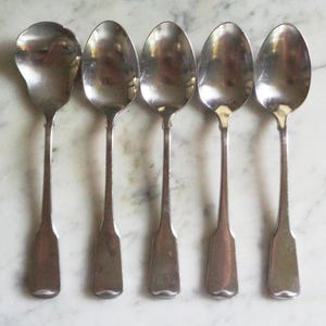 May include: A set of five silver-toned metal spoons arranged in a row. The spoons feature a smooth, reflective finish with a classic teardrop-shaped bowl and a flat, slightly flared handle end. Each spoon has a consistent design and metallic sheen.