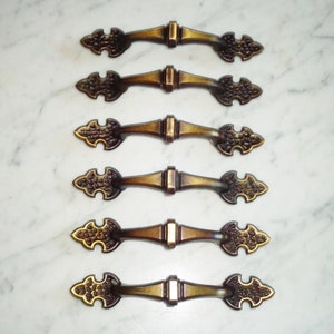 Könnte beinhalten: Satz von sechs antiken goldfarbenen Schrank-Schubladengriffen mit verzierten, detaillierten Designs. Jeder Griff hat ein dekoratives, spitzes Ende.