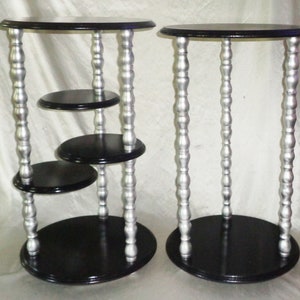 Pode incluir: Duas mesas de apoio de vários níveis, pretas e prateadas, com tampos redondos e várias prateleiras. As mesas têm um acabamento prateado decorativo nas pernas.