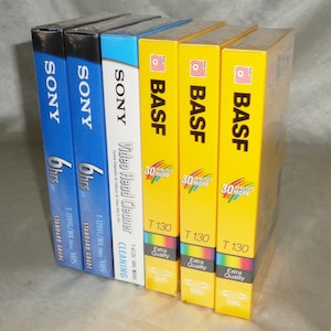 May include: A collection of VHS tapes, including brands like Sony and BASF. The tapes are in their original packaging, with labels indicating the tape length and type. The Sony tapes are blue, and the BASF tapes are yellow.