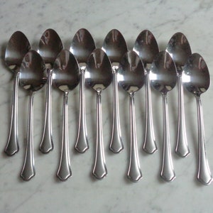May include: A set of twelve silver spoons with ornate handles. The spoons are arranged in a row, with the handles pointing towards the viewer.