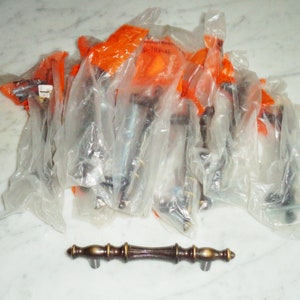 Puede incluir: Un paquete de 12 pomos de armario de latón antiguo con un diseño acanalado. Los pomos están empaquetados individualmente en bolsas de plástico transparentes con etiquetas naranjas.