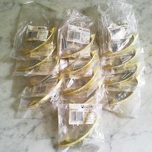 May include: A collection of ten gold-toned metal cabinet pulls, each individually packaged in a clear plastic bag with an Amerock label. The curved handles feature a smooth finish and are accompanied by small metal mounting screws inside the bags.