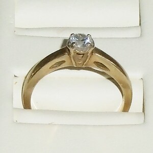 May include: A gold-colored engagement ring with a single, clear gemstone. The ring has a classic design with a band that tapers towards the center, where the gemstone is set. The ring is displayed in a white jewelry box.