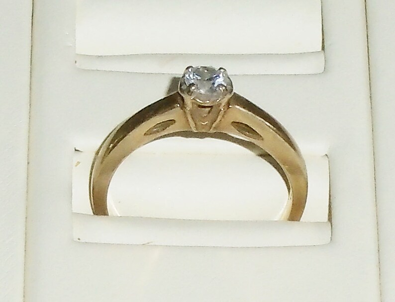 May include: A gold-colored engagement ring with a single, clear gemstone. The ring has a classic design with a band that tapers towards the center, where the gemstone is set. The ring is displayed in a white jewelry box.