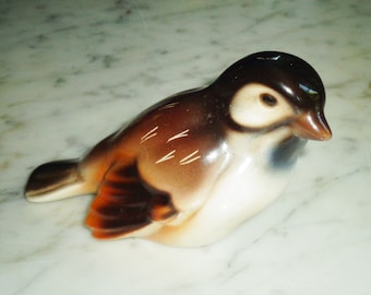 May include: A ceramic figurine of a brown and white bird with a black head. The bird is sitting on a white marble surface.