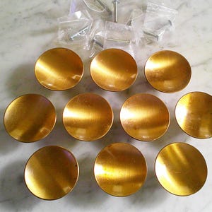 May include: A set of nine circular, concave cabinet knobs with a brushed gold finish, arranged in three rows. Each knob features a smooth, reflective surface. Included are nine small, clear plastic bags containing metal mounting screws for installation.
