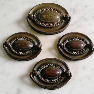May include: Four antique bronze drawer pulls. Each pull has a circular base with an oval center and a decorative beaded edge. The pulls have a dark brown finish and small, rounded handles on either side of the oval center.