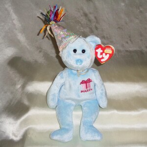 May include: A light blue Ty Beanie Baby bear wearing a party hat with colorful streamers. The bear has black eyes, a blue nose, and a gift graphic with the word "March" on its chest. A red heart-shaped tag is attached.