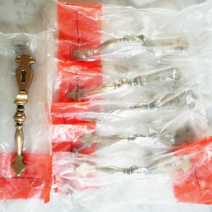 Op de afbeelding: Een set van zes antieke deurgrepen van goudkleurig metaal, verpakt in doorzichtige plastic zakken met rode labels. De grepen hebben een sleutelgat en een sierlijk ontwerp.