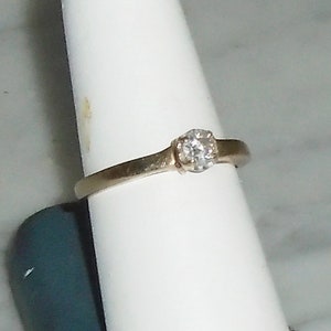 May include: A gold-toned ring with a single, clear diamond set in a prong setting. The band is thin and smooth, and the diamond is round. The ring is displayed on a white surface.
