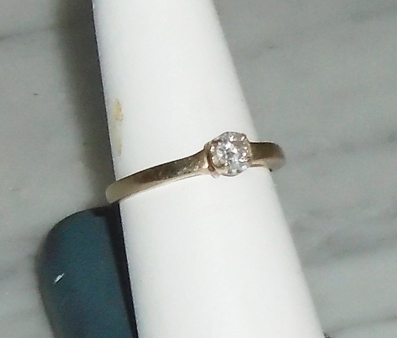 May include: A gold-toned ring with a single, clear diamond set in a prong setting. The band is thin and smooth, and the diamond is round. The ring is displayed on a white surface.