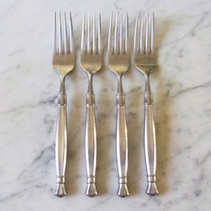 May include: A set of four silver-toned metal forks arranged vertically. Each fork features a four-tined head and a slender, tapered handle with a distinct central ridge and a flared, rounded base. The metal has a smooth, polished finish.