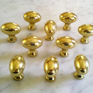 Könnte beinhalten: Eine Sammlung von zehn glänzenden, goldfarbenen Schrankknöpfen. Die Knöpfe haben eine abgerundete, ovale Form mit einem kurzen, dekorativen Sockel. Sie sind auf einer weißen und grauen Marmoroberfläche angeordnet und reflektieren das Licht.