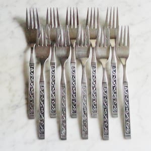 May include: A set of ten stainless steel forks arranged in a vertical row. Each fork features a long, slender handle decorated with a repeating dark, recessed scrollwork pattern that runs down the centre of the stem, ending in a flat base.