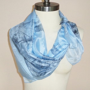 May include: Light blue silk scarf featuring a detailed illustration of a sailing ship. The scarf is draped around a mannequin, showcasing its flowing, lightweight fabric. The design is a classic nautical theme, perfect for adding a touch of elegance to any outfit.