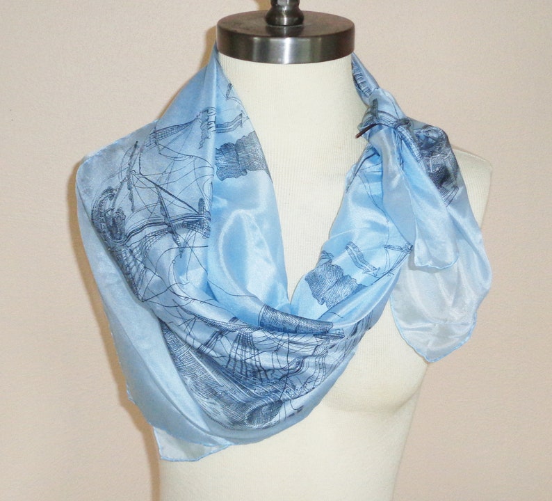 May include: Light blue silk scarf featuring a detailed illustration of a sailing ship. The scarf is draped around a mannequin, showcasing its flowing, lightweight fabric. The design is a classic nautical theme, perfect for adding a touch of elegance to any outfit.