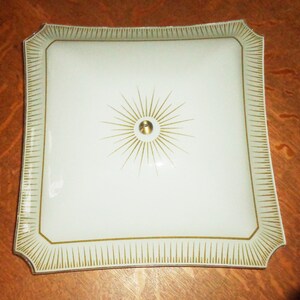 May include: A white square ceiling light fixture with a gold starburst design and a gold trim.