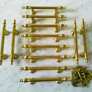 Puede incluir: Un conjunto de herrajes dorados para armarios, que incluye varios tiradores horizontales largos y tiradores verticales. Los herrajes están dispuestos sobre una superficie de mármol blanco y gris, con un pequeño montón de tornillos a juego.