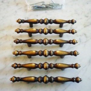 May include: Set of six antique brass cabinet knobs with a decorative design. The knobs are arranged in a row on a white marble surface.