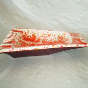 May include: A white ceramic ashtray with a red, green, and white splatter paint design. The ashtray has a rectangular shape with a slight curve on the bottom.