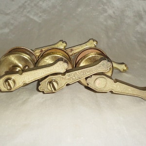 以下が含まれることがあります： 装飾的なデザインが施されたアンティークな真鍮製のドアノブが3つ。ノブは積み重ねられており、上のノブは他の2つのノブによって一部隠されています。