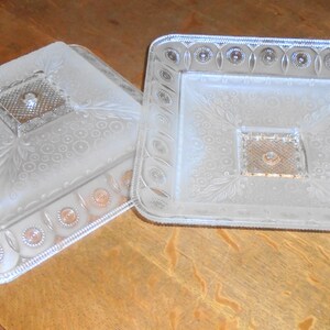 May include: Two clear glass serving dishes with a raised, embossed design. The dishes are square and have a frosted finish.