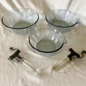 Può includere: Tre ciotole di vetro trasparente con bordi blu e due manici in vetro e metallo. Le ciotole sono su una superficie bianca. Ci sono due manici in vetro e metallo con una parte in metallo nero.